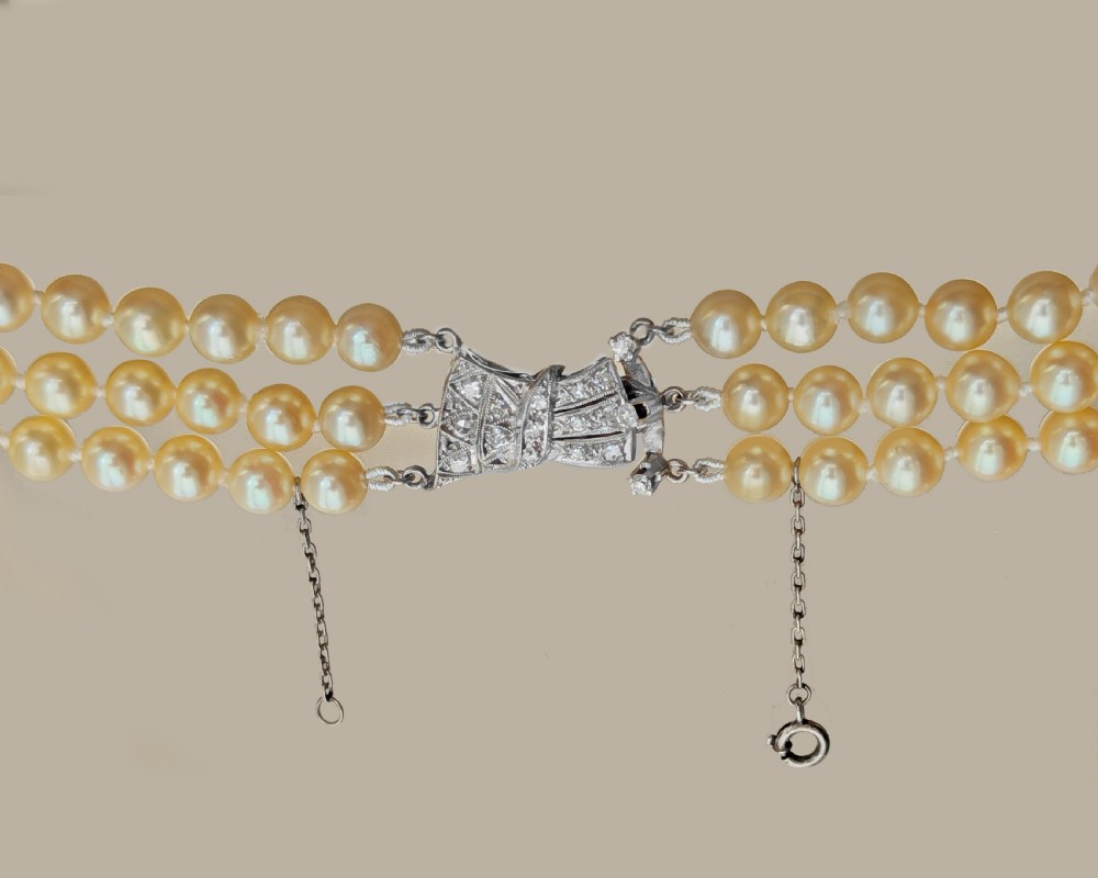 A 1950s triple strand cream pearl necklace featuring an 18ct white gold bow clasp set with brilliant diamonds. View IV