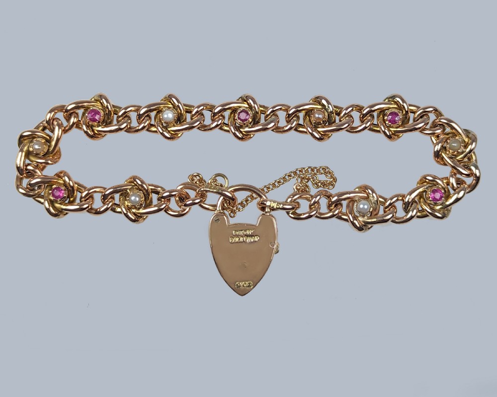A Victorian 9ct yellow gold knot link chain bracelet featuring embedded rubies and seed pearls, secured by a heart-shaped padlock clasp with a safety chain. View VI