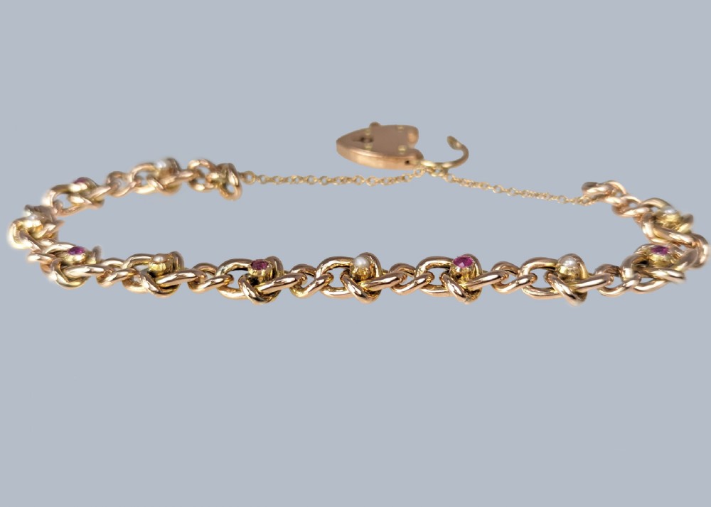 A Victorian 9ct yellow gold knot link chain bracelet featuring embedded rubies and seed pearls, secured by a heart-shaped padlock clasp with a safety chain. View IX