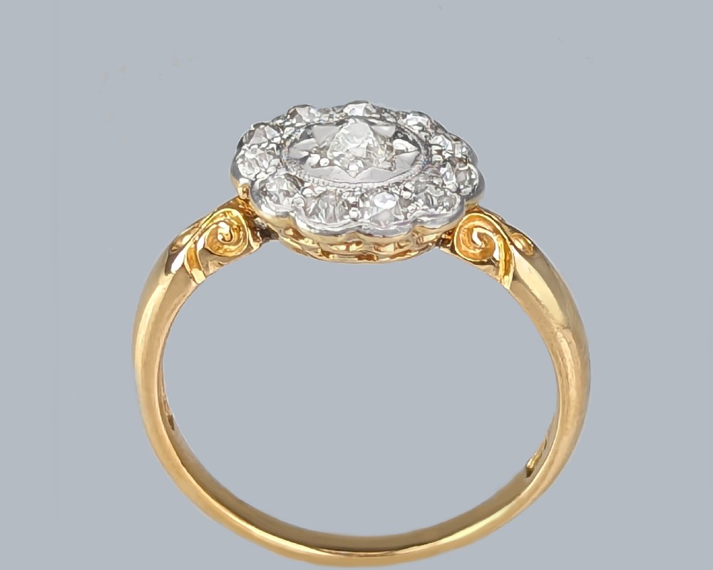 An Edwardian cluster ring with a central old cut diamond surrounded by smaller accent diamonds in a circular setting, set on an 18ct yellow gold band with carved shoulders. View III