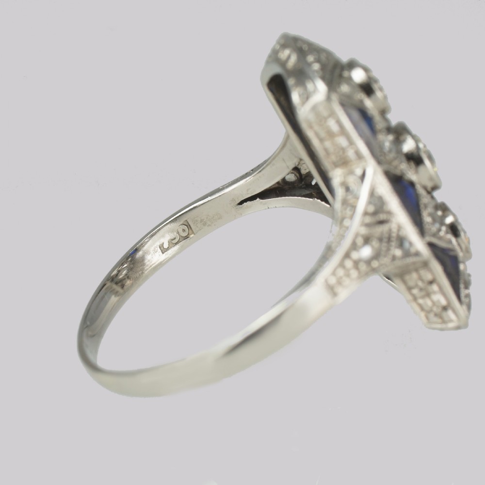 An Art Deco style rectangular plaque ring featuring three central round diamonds flanked by millegrain detail and geometric blue sapphire accents set in silver-toned metal. View IX
