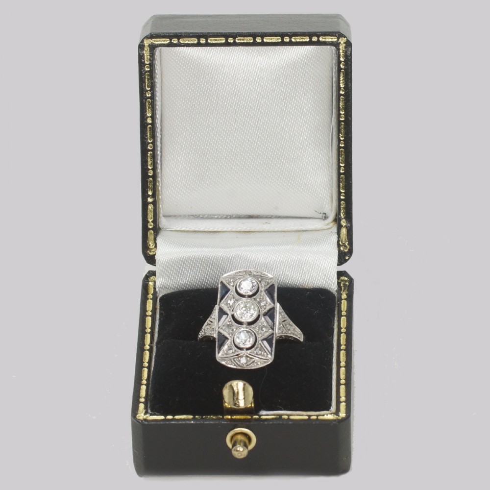 An Art Deco style rectangular plaque ring featuring three central round diamonds flanked by millegrain detail and geometric blue sapphire accents set in silver-toned metal. View XII