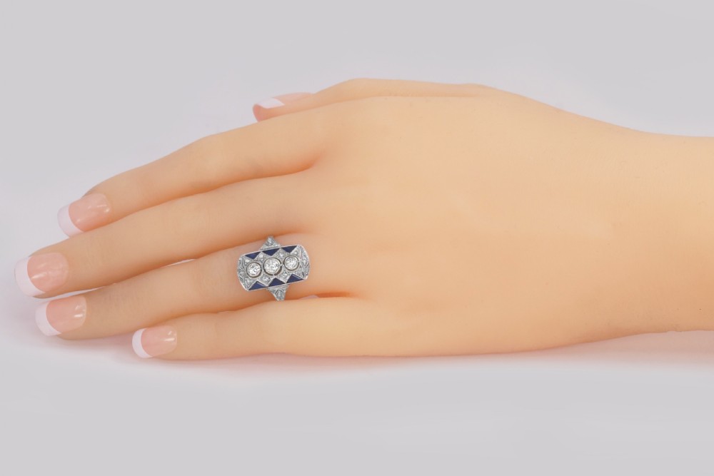 An Art Deco style rectangular plaque ring featuring three central round diamonds flanked by millegrain detail and geometric blue sapphire accents set in silver-toned metal. View XIII
