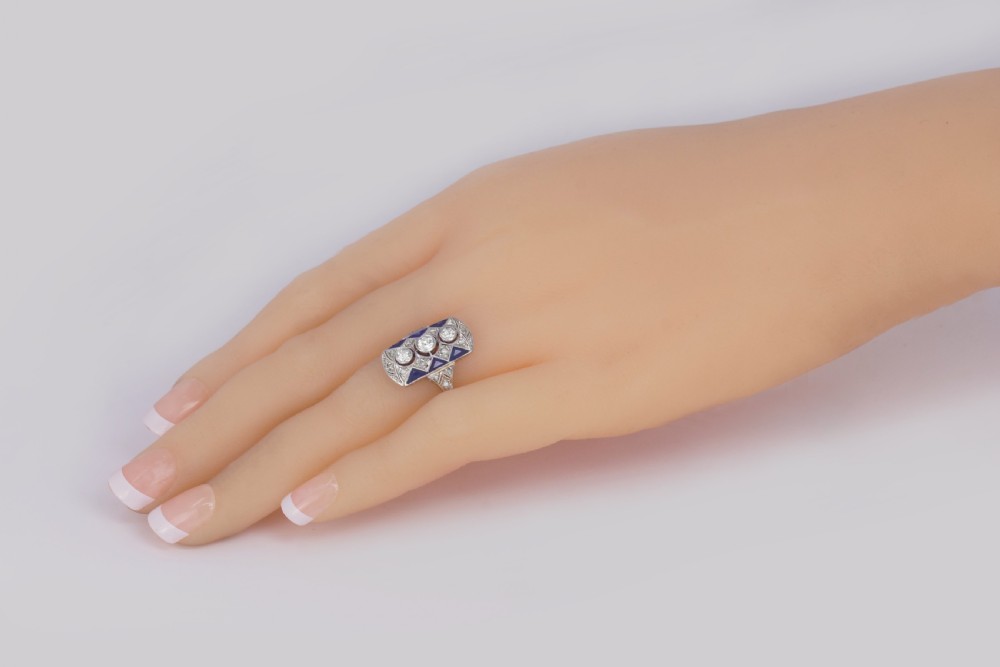 An Art Deco style rectangular plaque ring featuring three central round diamonds flanked by millegrain detail and geometric blue sapphire accents set in silver-toned metal. View XIV