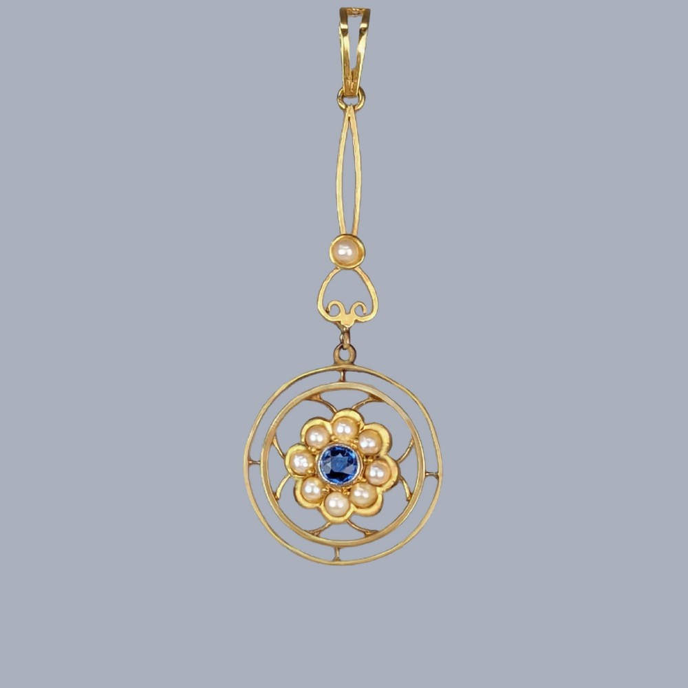 An Edwardian 15ct gold drop pendant featuring a central round blue sapphire surrounded by seed pearls within an openwork circular frame.