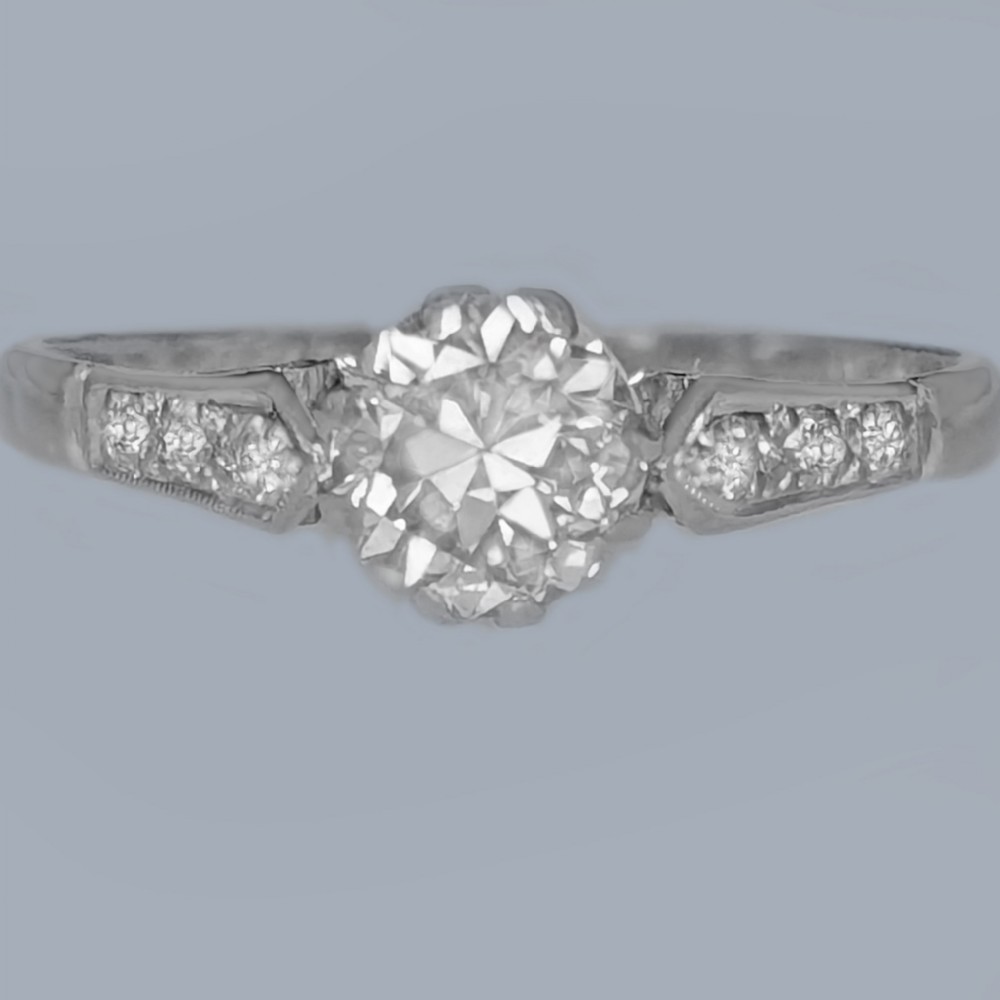 An Art Deco diamond solitaire ring featuring a round central diamond flanked by geometric shoulders set with small accent diamonds in white gold and platinum.