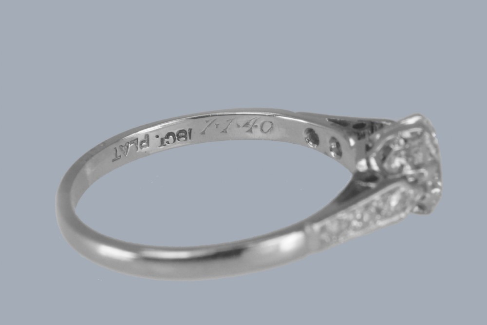 An Art Deco diamond solitaire ring featuring a round central diamond flanked by geometric shoulders set with small accent diamonds in white gold and platinum. View VII