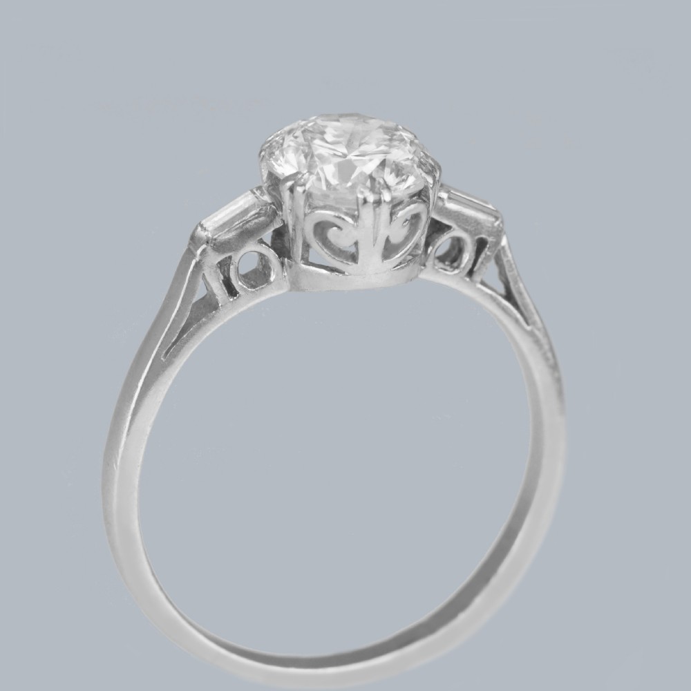An Art Deco platinum solitaire ring featuring a round brilliant cut diamond flanked by stepped baguette cut shoulder stones. View IV