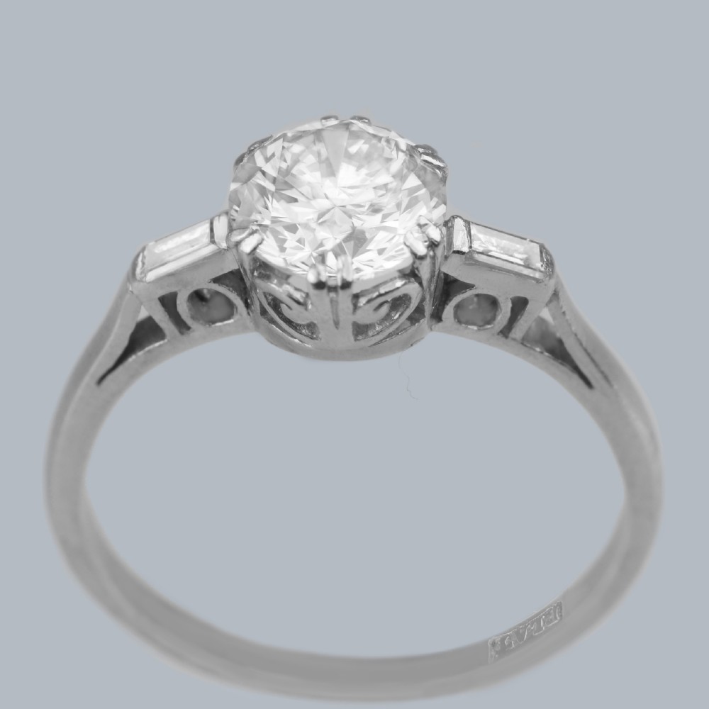 An Art Deco platinum solitaire ring featuring a round brilliant cut diamond flanked by stepped baguette cut shoulder stones. View IX