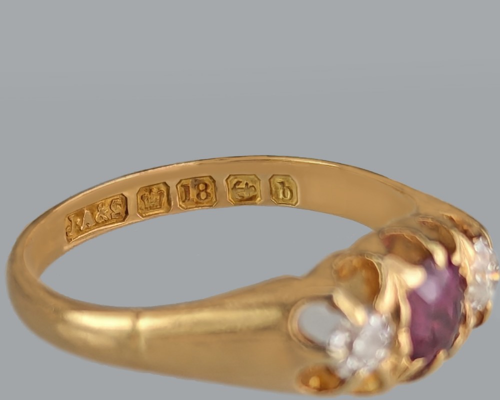 An antique 18ct yellow gold Edwardian ring featuring a central oval cut ruby flanked by two round diamonds set into carved scrollwork shoulders. View VII