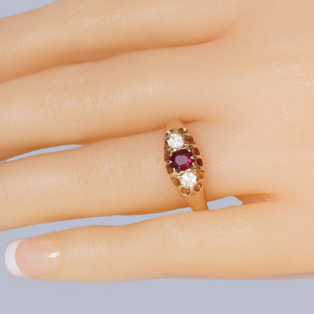 An antique 18ct yellow gold Edwardian ring featuring a central oval cut ruby flanked by two round diamonds set into carved scrollwork shoulders. View VIII