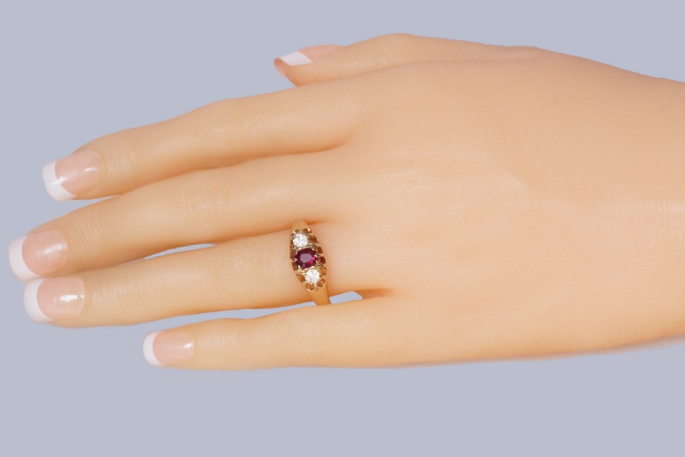 An antique 18ct yellow gold Edwardian ring featuring a central oval cut ruby flanked by two round diamonds set into carved scrollwork shoulders. View XV