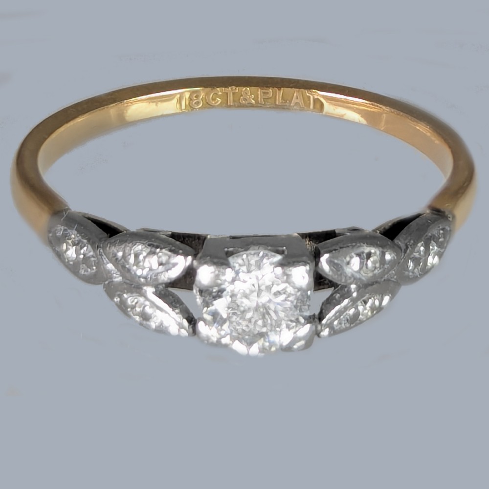 An Edwardian solitaire ring featuring a central round diamond in a platinum setting with diamond leaf shoulders on an 18ct yellow gold band. View V