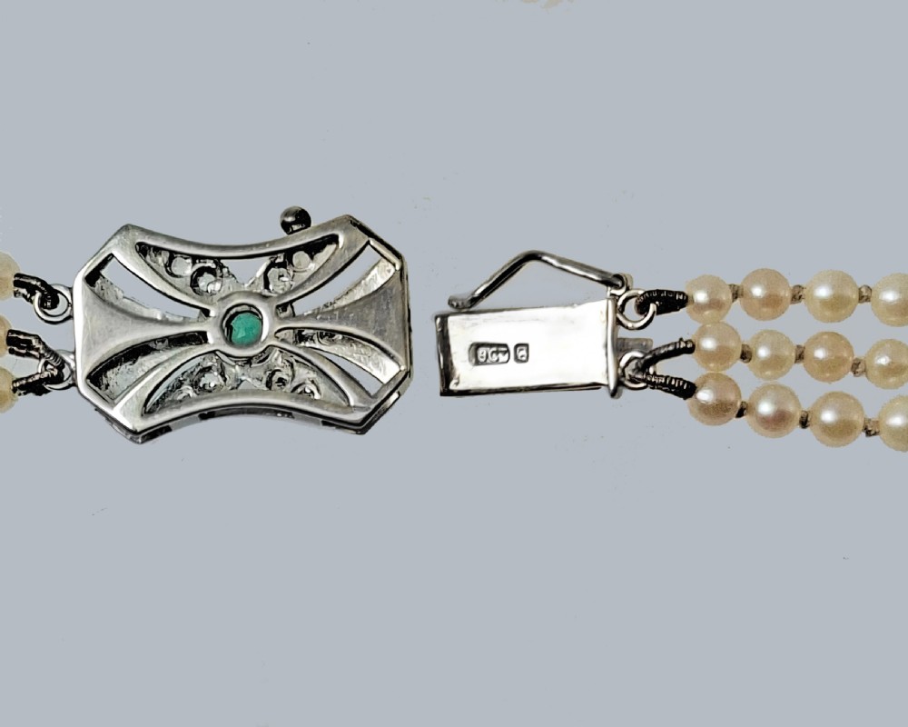 A 1950s three-strand pearl necklace featuring graduated pearls joined by a geometric clasp set with diamonds and a central green emerald. View IV