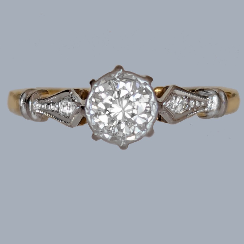 An antique 1920s diamond solitaire ring with a round cut central diamond set in platinum with geometric shoulders on an 18ct yellow gold band.