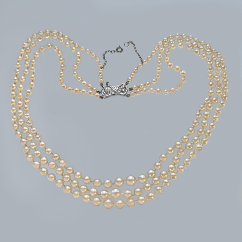 A 1950s triple strand pearl necklace featuring graduated cream pearls and a geometric diamond-set clasp with a safety chain.