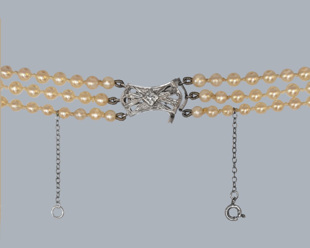 A 1950s triple strand pearl necklace featuring graduated cream pearls and a geometric diamond-set clasp with a safety chain. View V