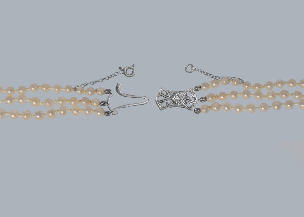 A 1950s triple strand pearl necklace featuring graduated cream pearls and a geometric diamond-set clasp with a safety chain. View VI