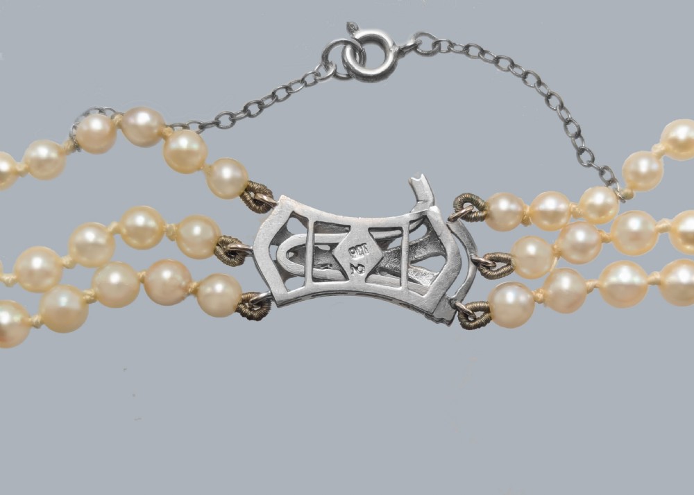 A 1950s triple strand pearl necklace featuring graduated cream pearls and a geometric diamond-set clasp with a safety chain. View VII