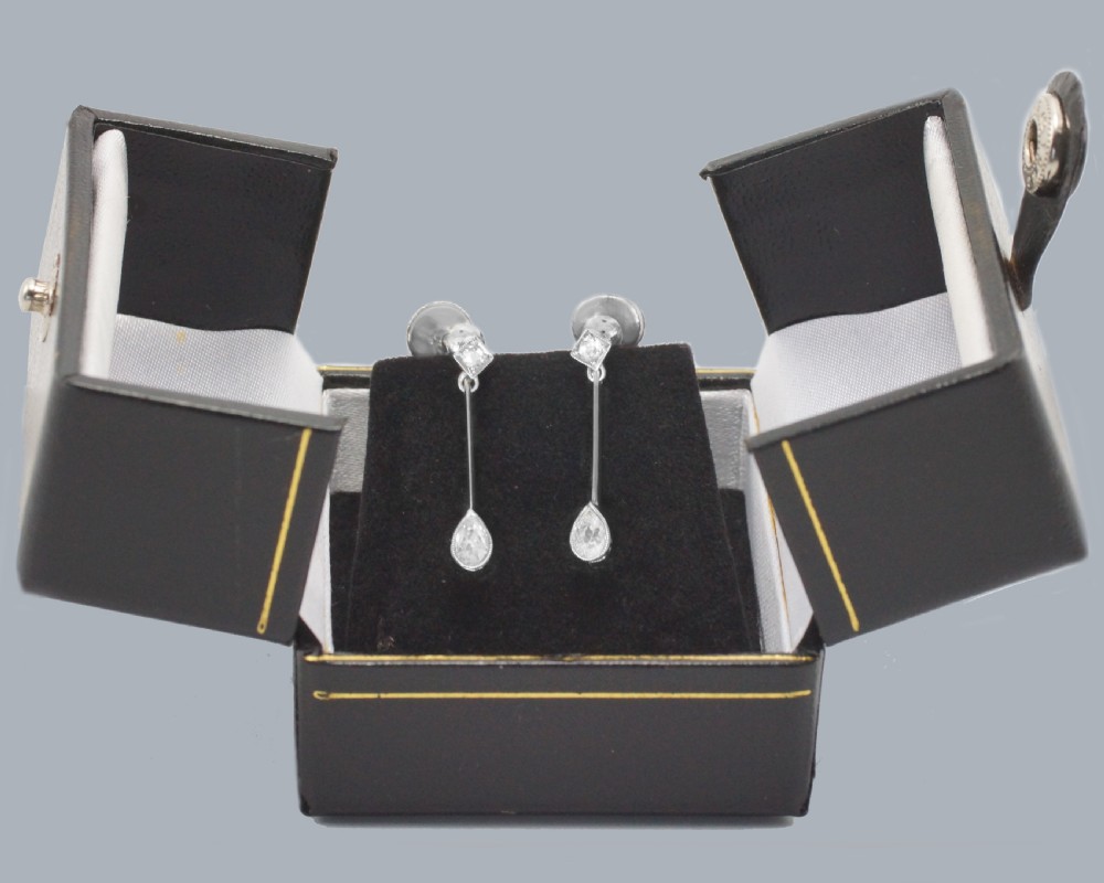 A pair of Edwardian 18ct white gold drop earrings featuring pear-shaped diamonds suspended from linear knife-edge bars and diamond-shaped top studs. View VI