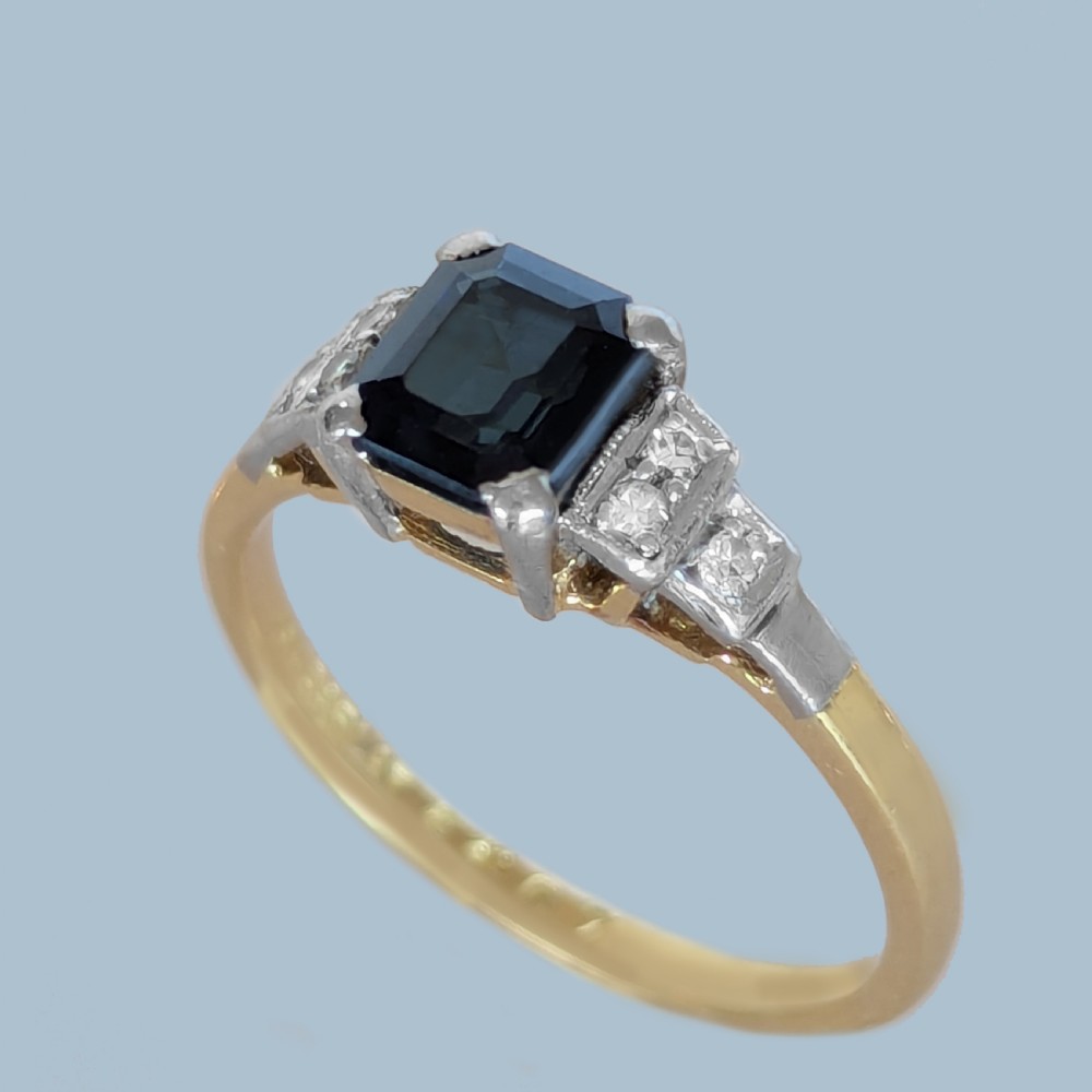 An Art Deco ring featuring a central emerald-cut deep blue sapphire flanked by geometric stepped platinum shoulders set with small diamonds on an 18ct yellow gold band. View IV
