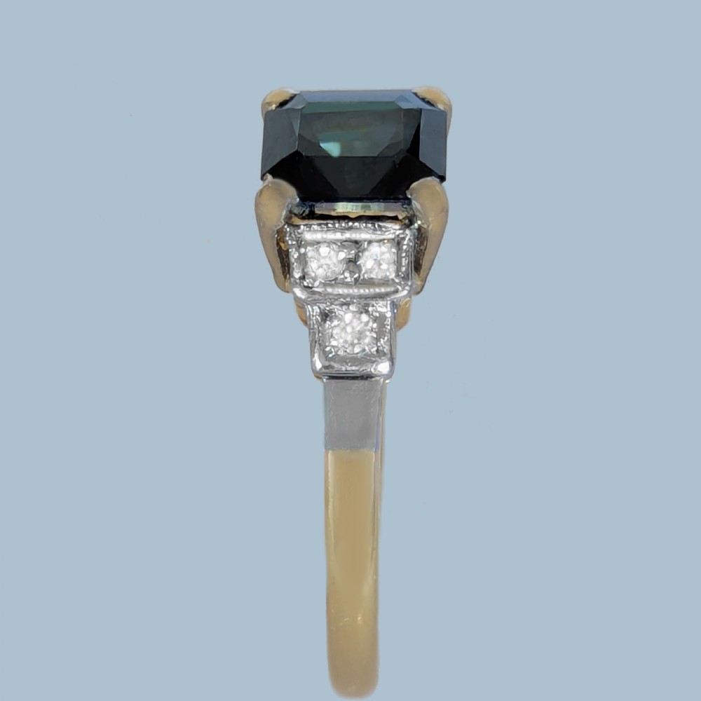 An Art Deco ring featuring a central emerald-cut deep blue sapphire flanked by geometric stepped platinum shoulders set with small diamonds on an 18ct yellow gold band. View VII