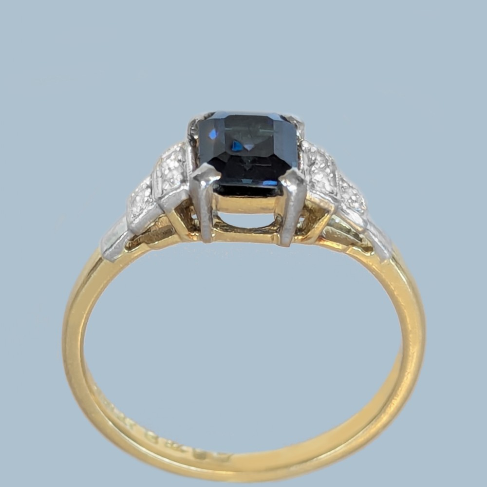 An Art Deco ring featuring a central emerald-cut deep blue sapphire flanked by geometric stepped platinum shoulders set with small diamonds on an 18ct yellow gold band. View VIII