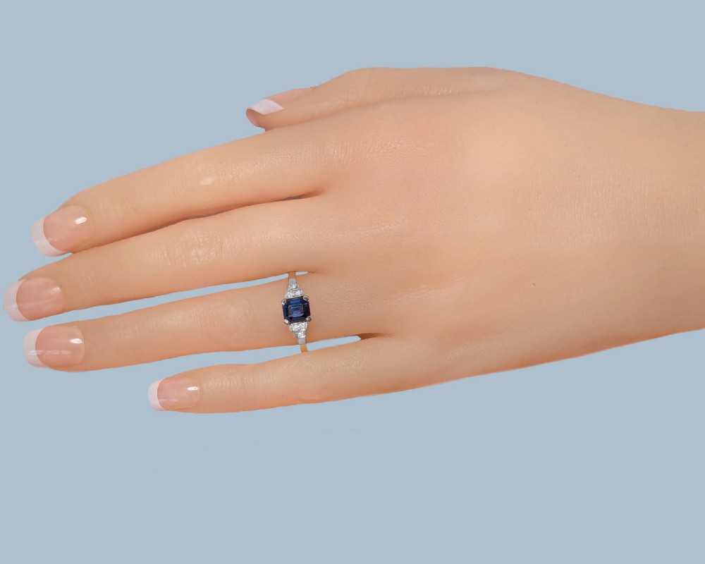 An Art Deco ring featuring a central emerald-cut deep blue sapphire flanked by geometric stepped platinum shoulders set with small diamonds on an 18ct yellow gold band. View X