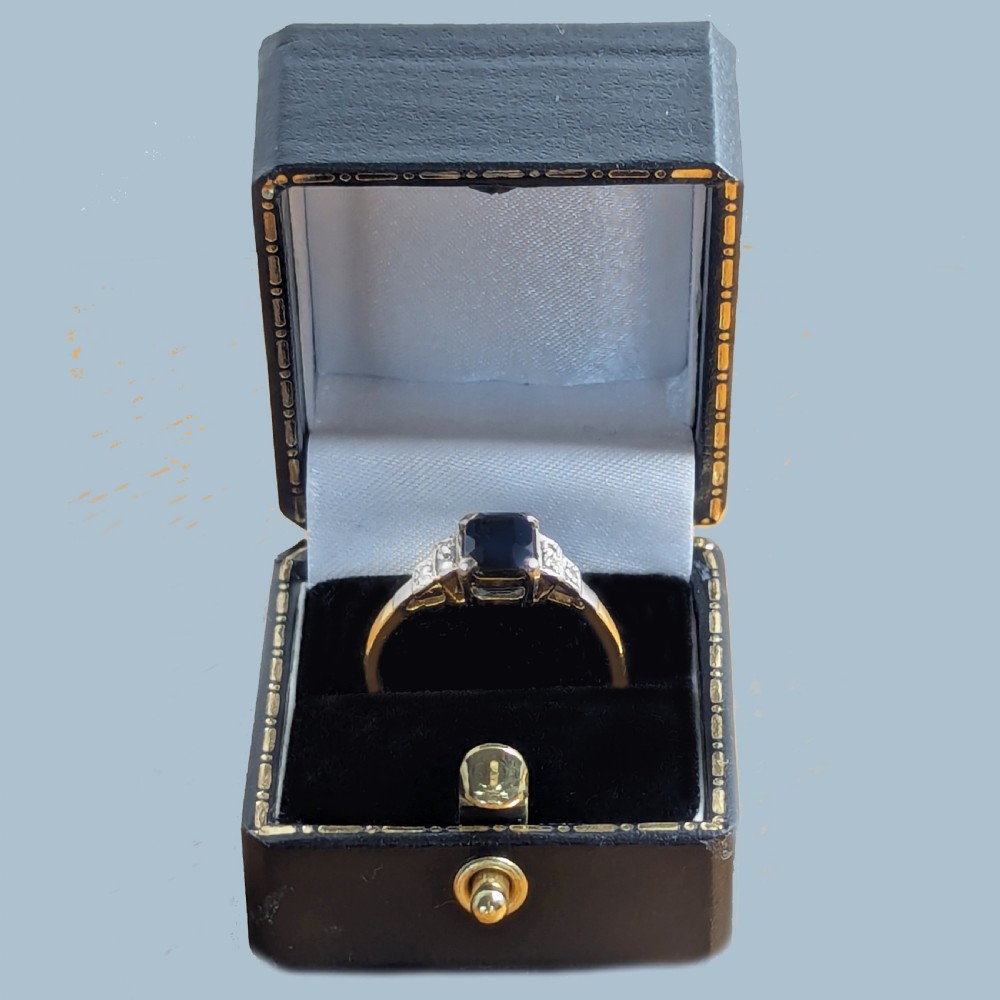 An Art Deco ring featuring a central emerald-cut deep blue sapphire flanked by geometric stepped platinum shoulders set with small diamonds on an 18ct yellow gold band. View XI