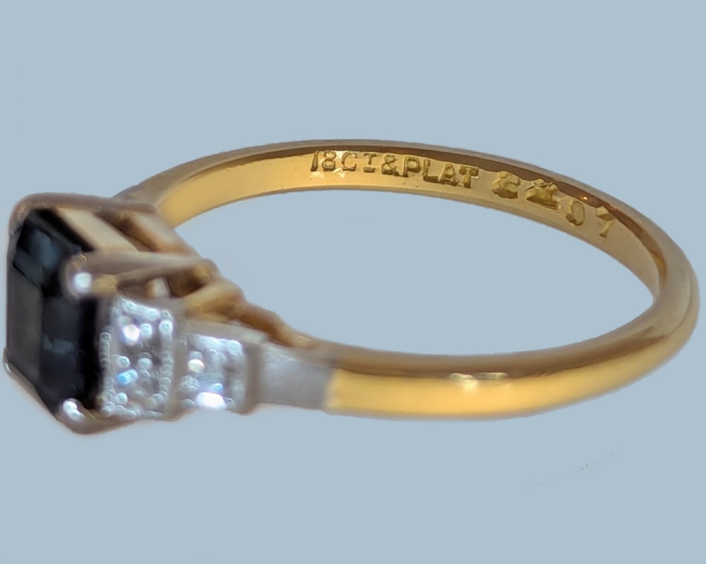 An Art Deco ring featuring a central emerald-cut deep blue sapphire flanked by geometric stepped platinum shoulders set with small diamonds on an 18ct yellow gold band. View XII