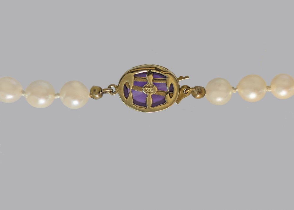 A single strand of uniform cream pearls featuring a central oval cabochon amethyst clasp set in gold, displayed against a black backdrop. View IV