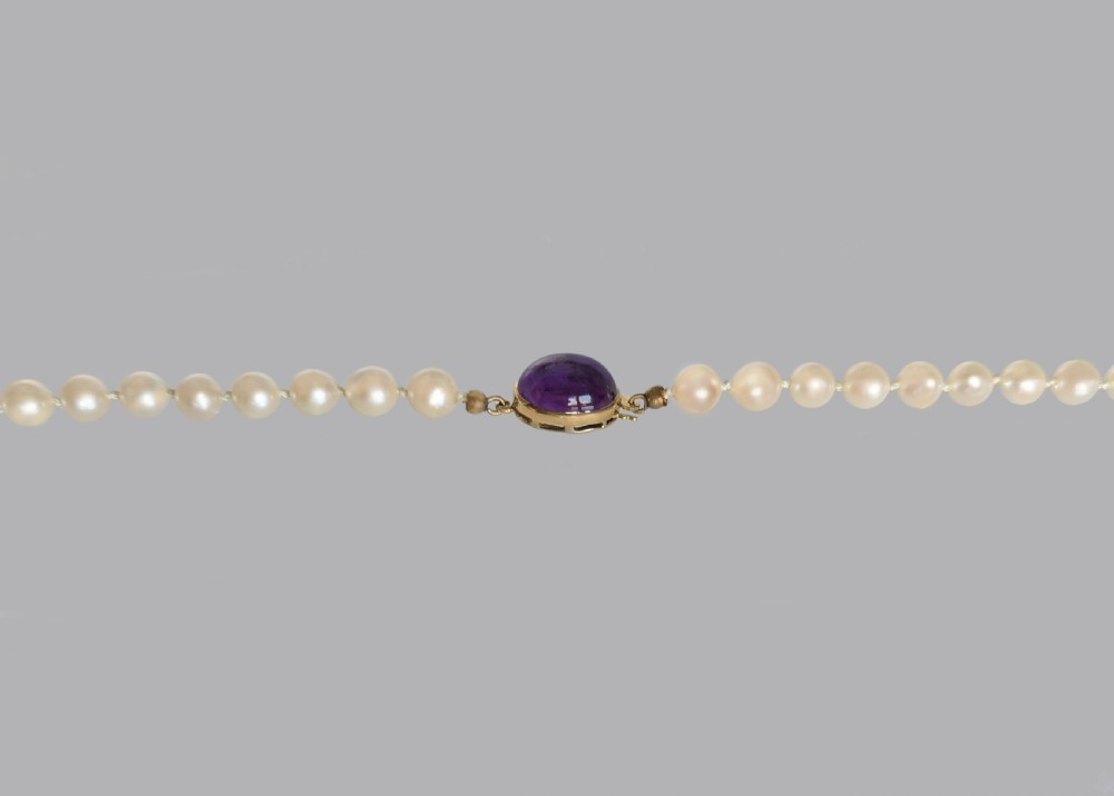 A single strand of uniform cream pearls featuring a central oval cabochon amethyst clasp set in gold, displayed against a black backdrop. View V