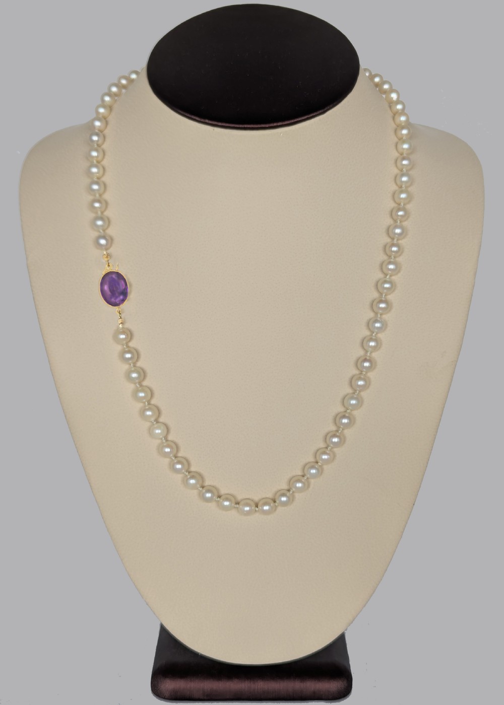 A single strand of uniform cream pearls featuring a central oval cabochon amethyst clasp set in gold, displayed against a black backdrop. View VII