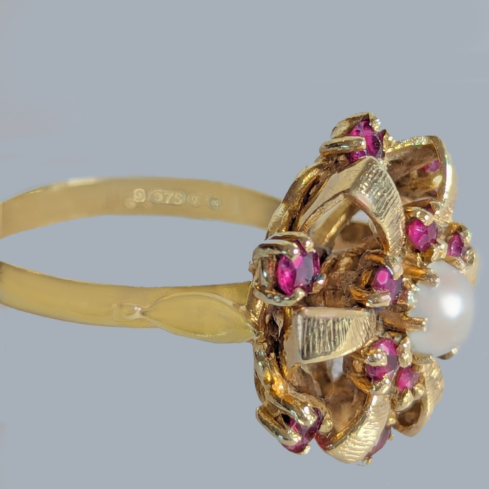 A 9ct yellow gold cluster ring featuring a central white pearl surrounded by textured gold ribbon loops set with round red rubies. View V