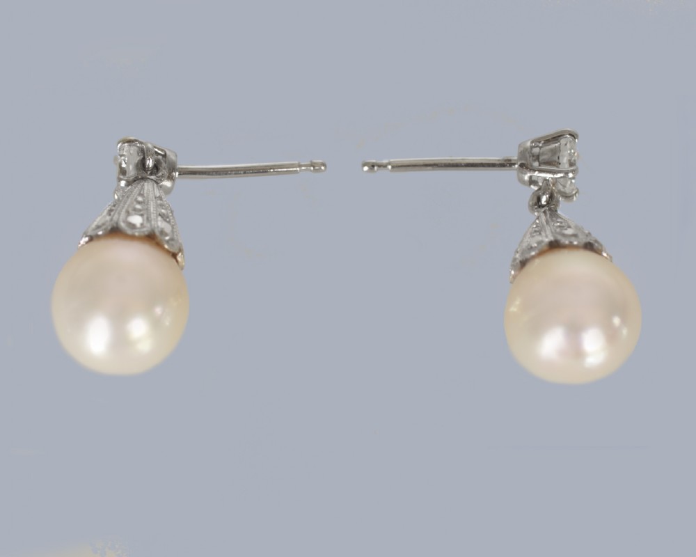 A pair of 1950s 18ct white gold drop earrings featuring round pearls suspended from fluted diamond-set caps and single brilliant-cut diamond studs. View V