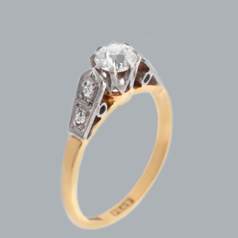 An Art Deco diamond solitaire ring featuring a round central stone set in platinum, flanked by geometric shoulders on an 18ct yellow gold band. View V