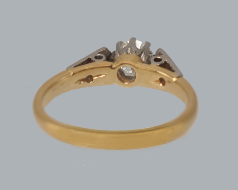 An Art Deco diamond solitaire ring featuring a round central stone set in platinum, flanked by geometric shoulders on an 18ct yellow gold band. View VI