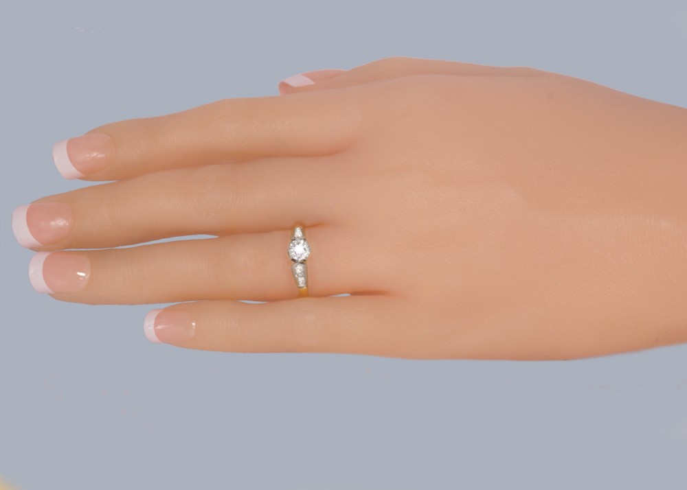 An Art Deco diamond solitaire ring featuring a round central stone set in platinum, flanked by geometric shoulders on an 18ct yellow gold band. View VIII