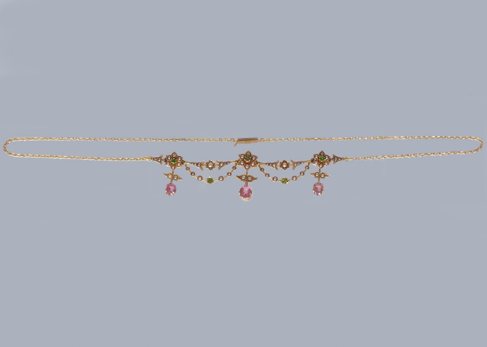 An Edwardian 18ct gold festoon necklace featuring delicate seed pearl floral motifs, small green garnets, and three oval pink sapphire drops. View VIII