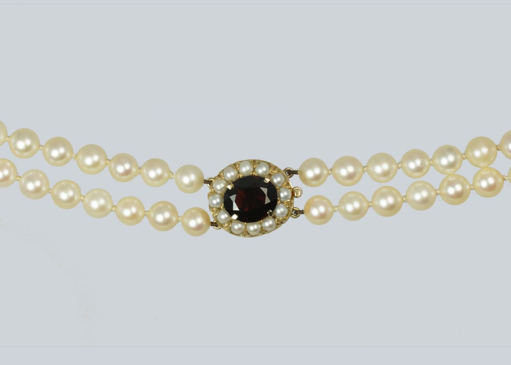A double strand cultured pearl necklace featuring uniform cream pearls fastened with a round 9ct gold clasp inset with a dark red garnet stone. View VI