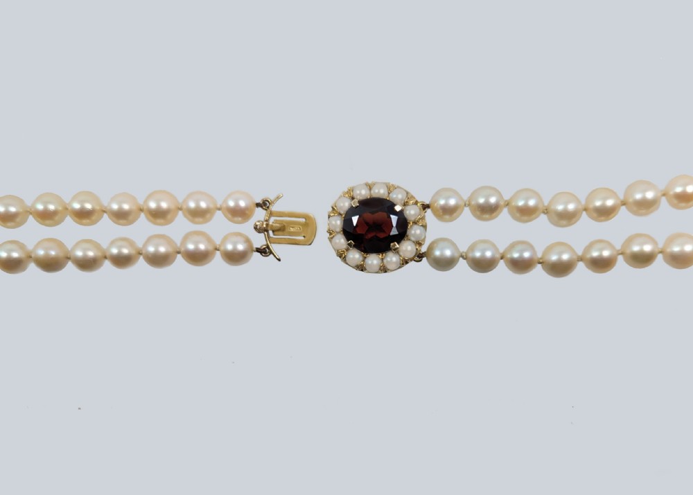 A double strand cultured pearl necklace featuring uniform cream pearls fastened with a round 9ct gold clasp inset with a dark red garnet stone. View VII