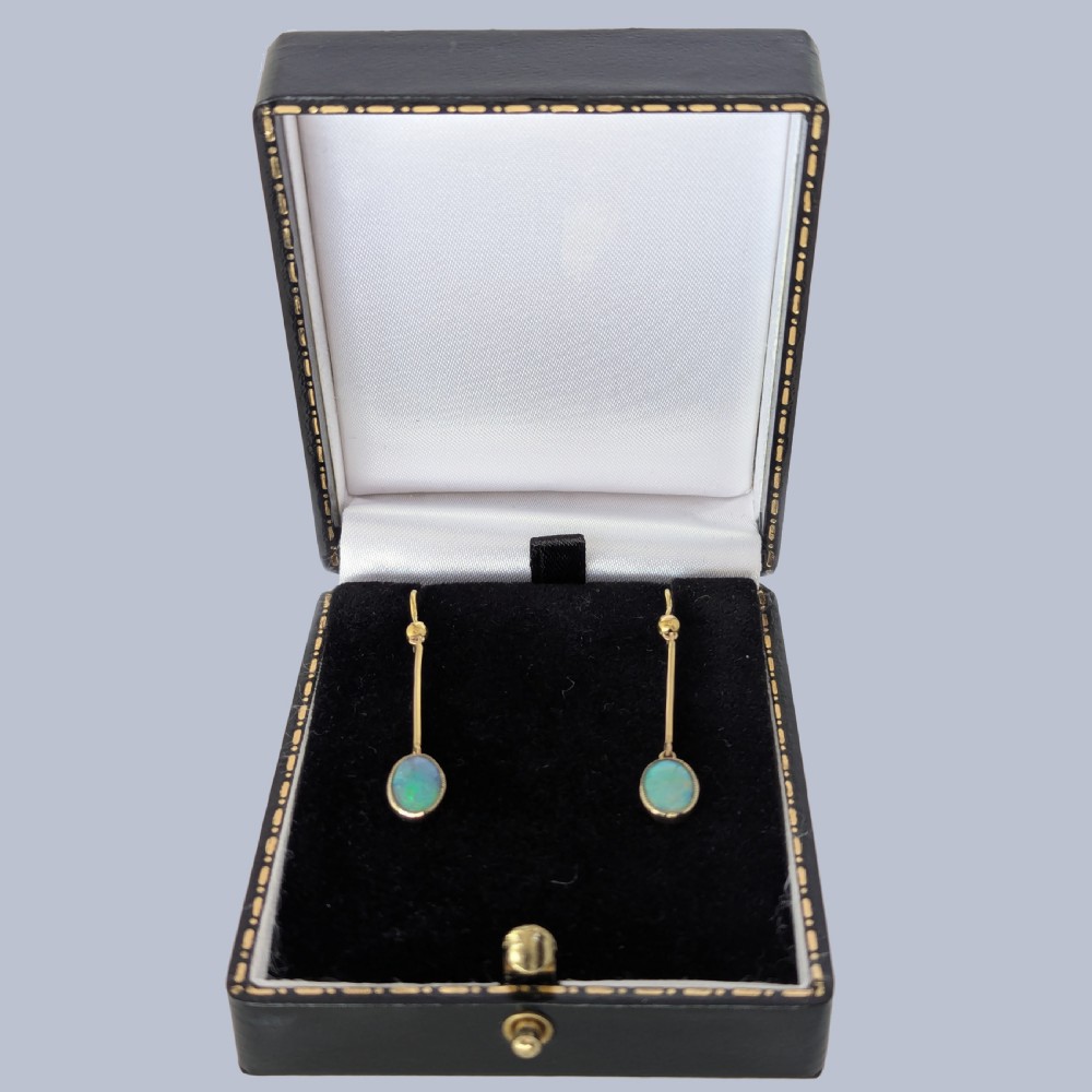 A pair of vintage 18 carat yellow gold drop earrings featuring slender vertical bars suspended from wire hooks and terminating in oval opal cabochons displaying blue and green play-of-colour within milgrain settings. View VI
