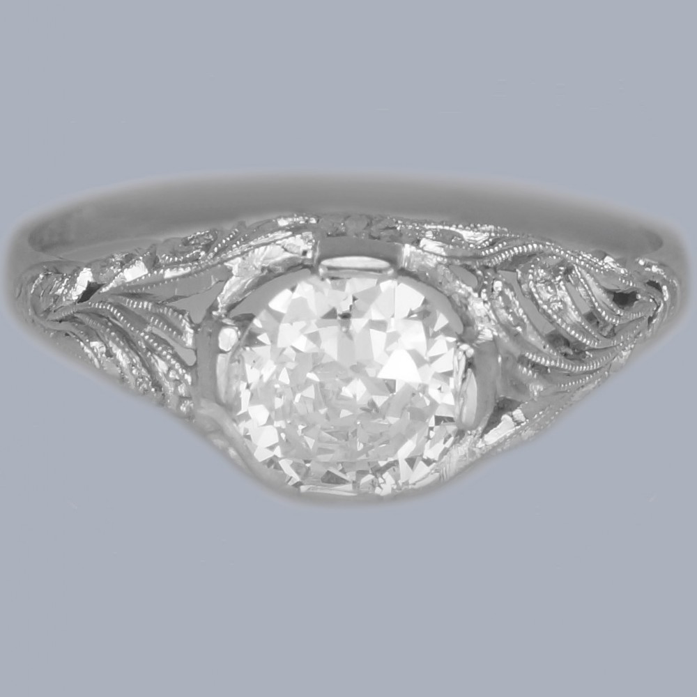 An Edwardian platinum engagement ring featuring a central round old cut diamond flanked by detailed filigree shoulders.