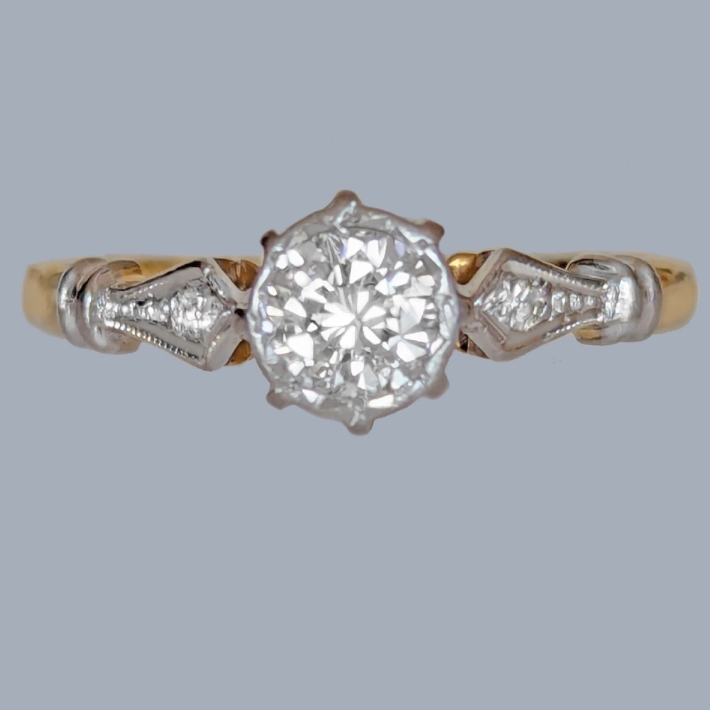 An antique 1920s diamond solitaire ring with a round cut central diamond set in platinum with geometric shoulders on an 18ct yellow gold band.