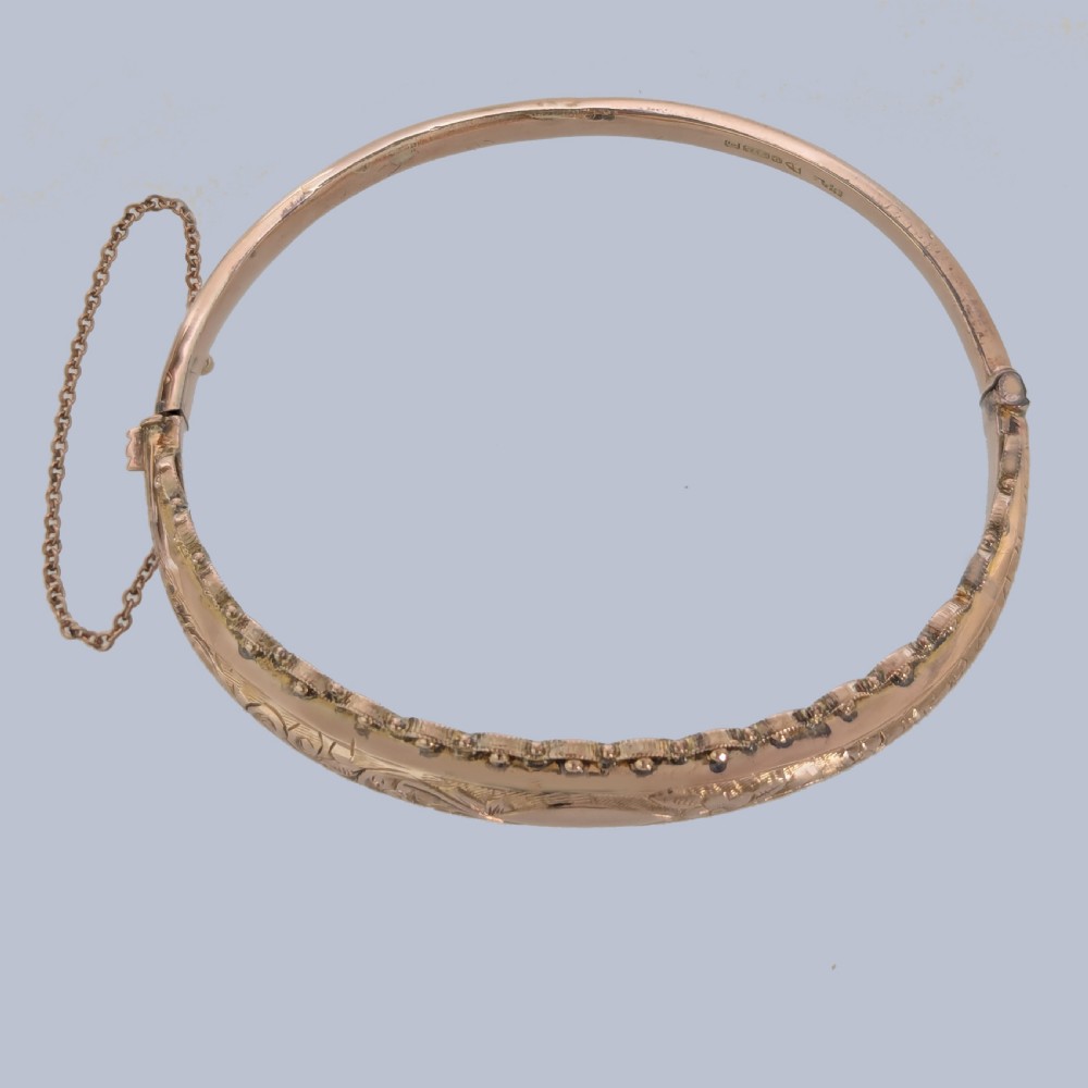 An Edwardian 9ct rose gold hinged bangle with foliate engraving, beaded border trim, and a safety chain. View VI