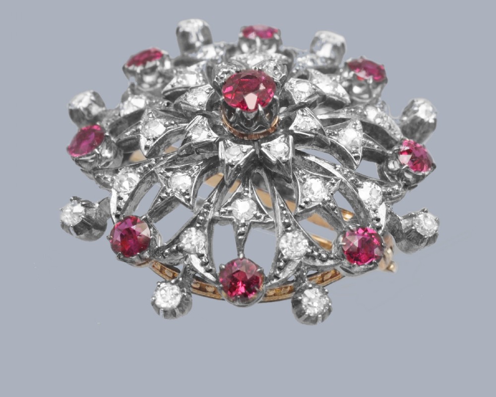 A circular Victorian brooch rendered in silver and gold, featuring a central round ruby surrounded by intricate diamond-set starburst openwork and an outer border of alternating rubies and diamonds. View V
