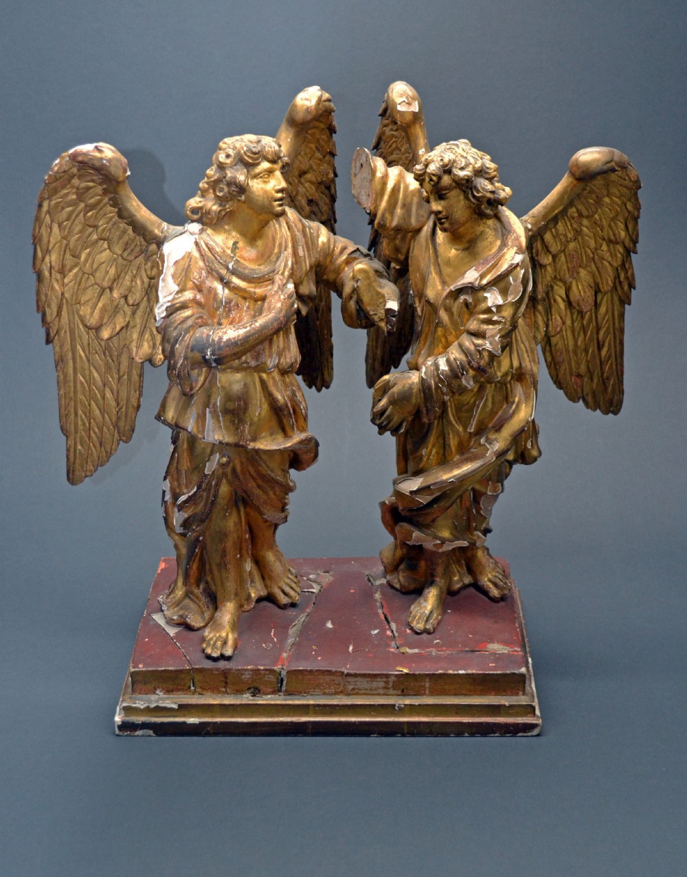 Two antique carved wooden figures of archangels with large wings, finished in distressed gold leaf and standing on a red-painted rectangular base.