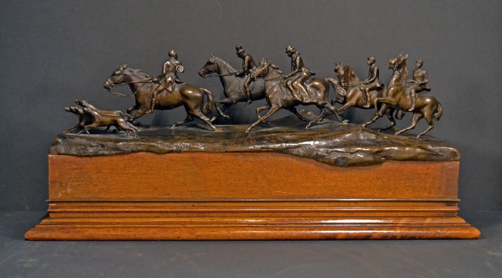 A detailed dark bronze sculpture depicting multiple riders on horseback following hunting hounds, mounted on a rectangular wooden base.