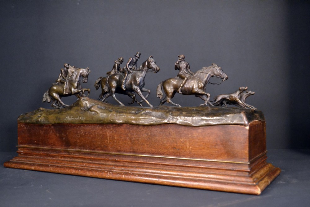 A detailed dark bronze sculpture depicting multiple riders on horseback following hunting hounds, mounted on a rectangular wooden base. View III