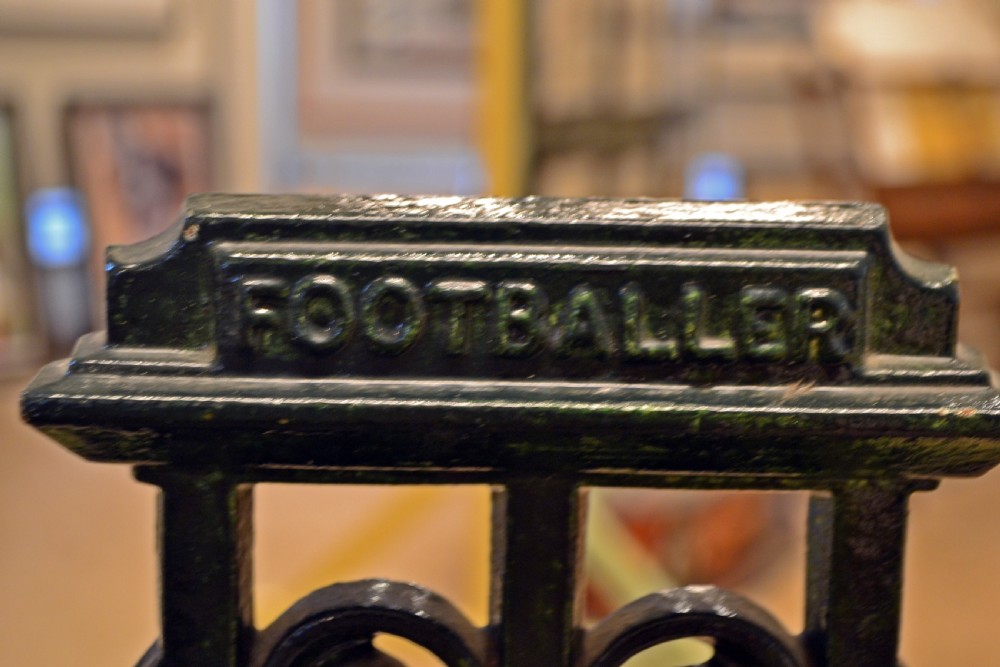 A black cast iron stick stand featuring a central relief figure of a nineteenth-century footballer holding a ball, framed by scrolling detail and twin drip trays. View II
